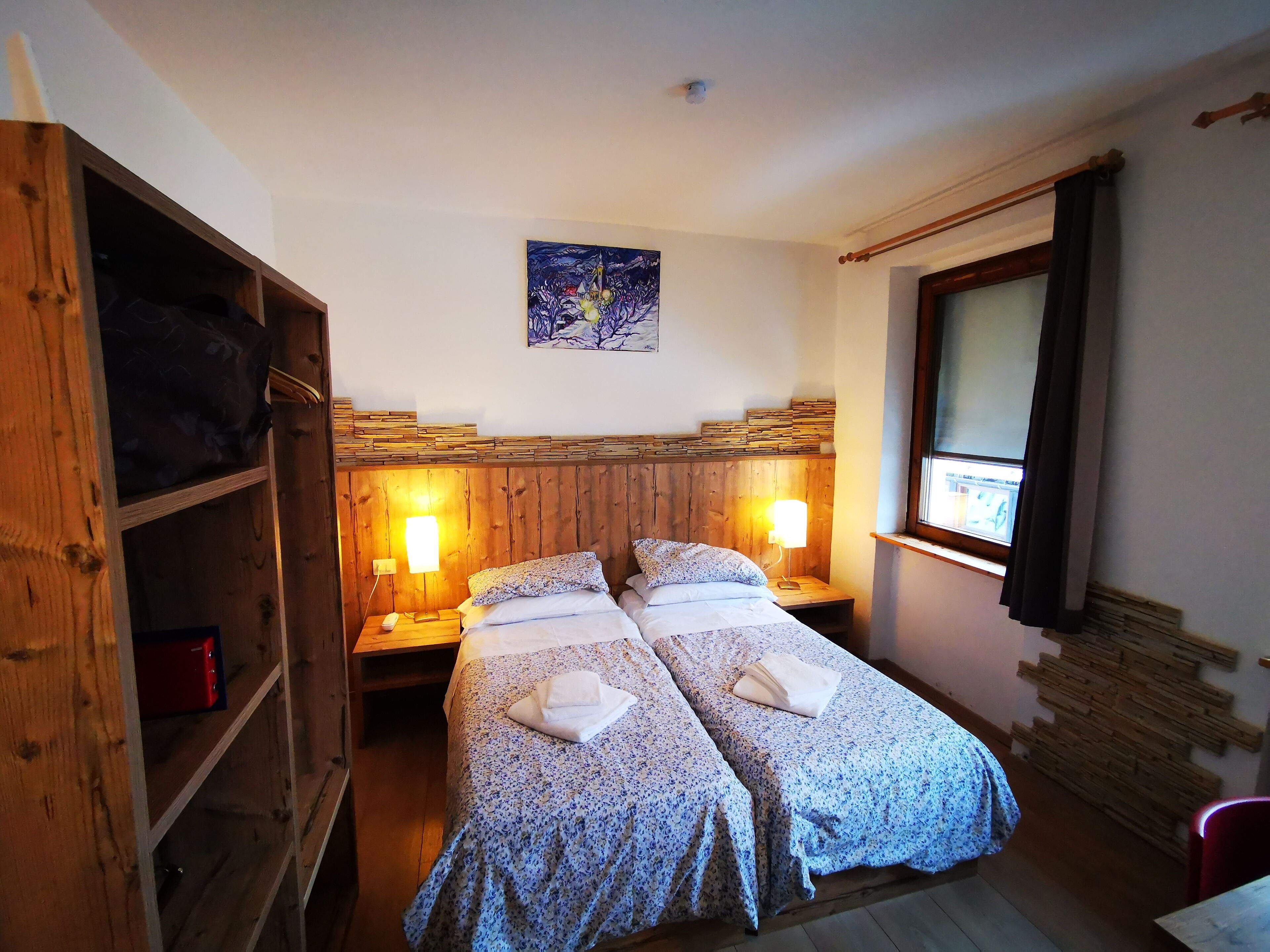 A room with two beds, wooden headboard, bedside lamps, a painting on the wall, and a window with curtains.