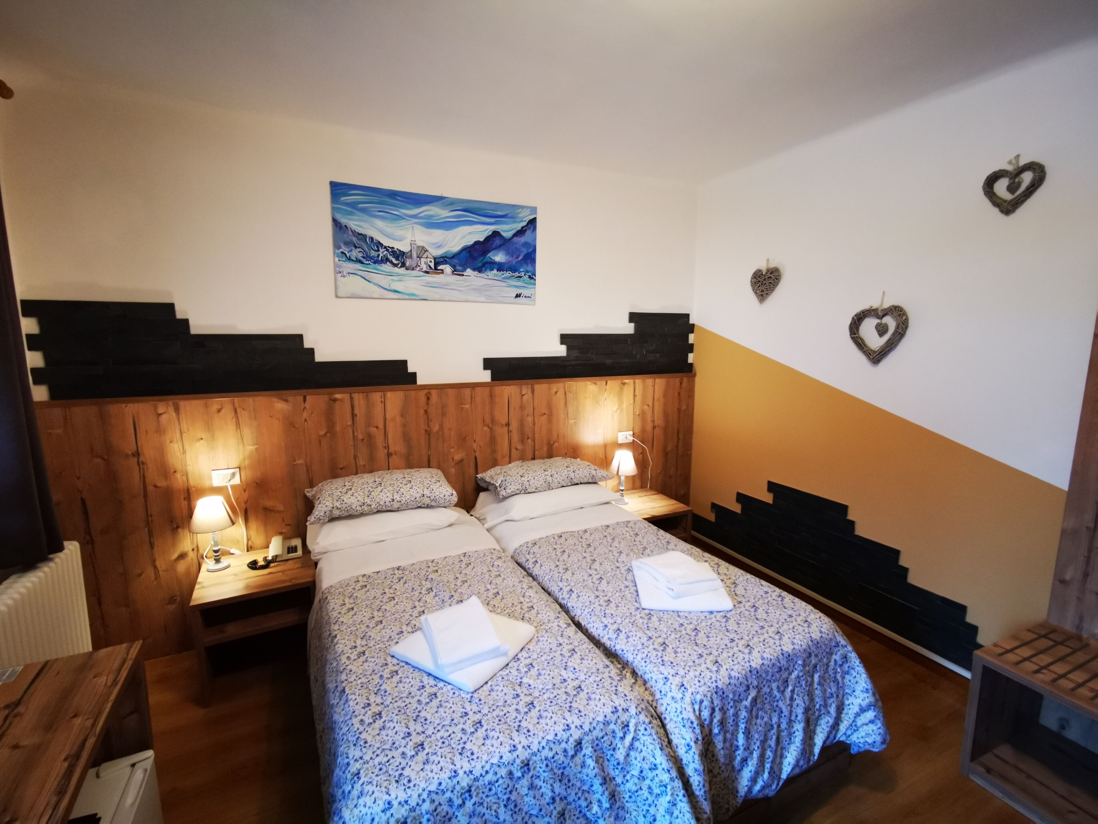A double room with two beds, wooden headboard, bedside lamps, a painting on the wall, and heart-shaped decorations.