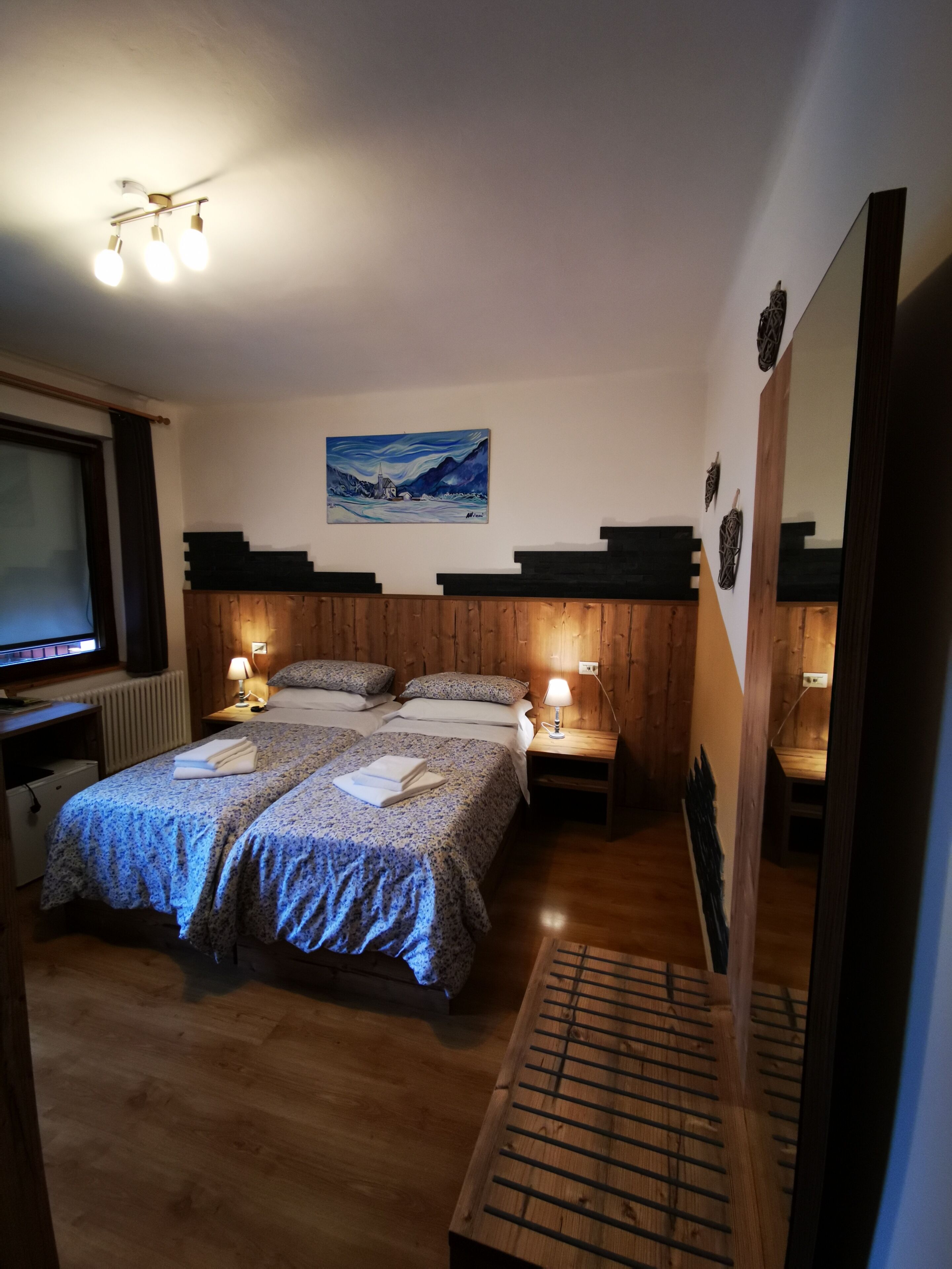 Un dormitorio con dos camas, un cuadro en la pared, cabeceros de madera, lámparas de noche, un banco de madera y un radiador.
