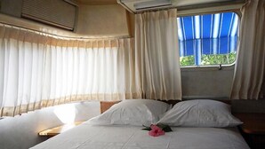 A bed with white linens and a single pink flower, a window with blue curtains, and an air conditioning unit above the window. - Airstream Paradise (Sainte-Anne)