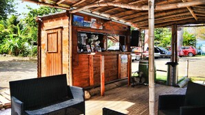 A wooden kiosk with a counter, surrounded by wicker chairs, and a covered area with a bamboo roof. - Airstream Paradise (Sainte-Anne)