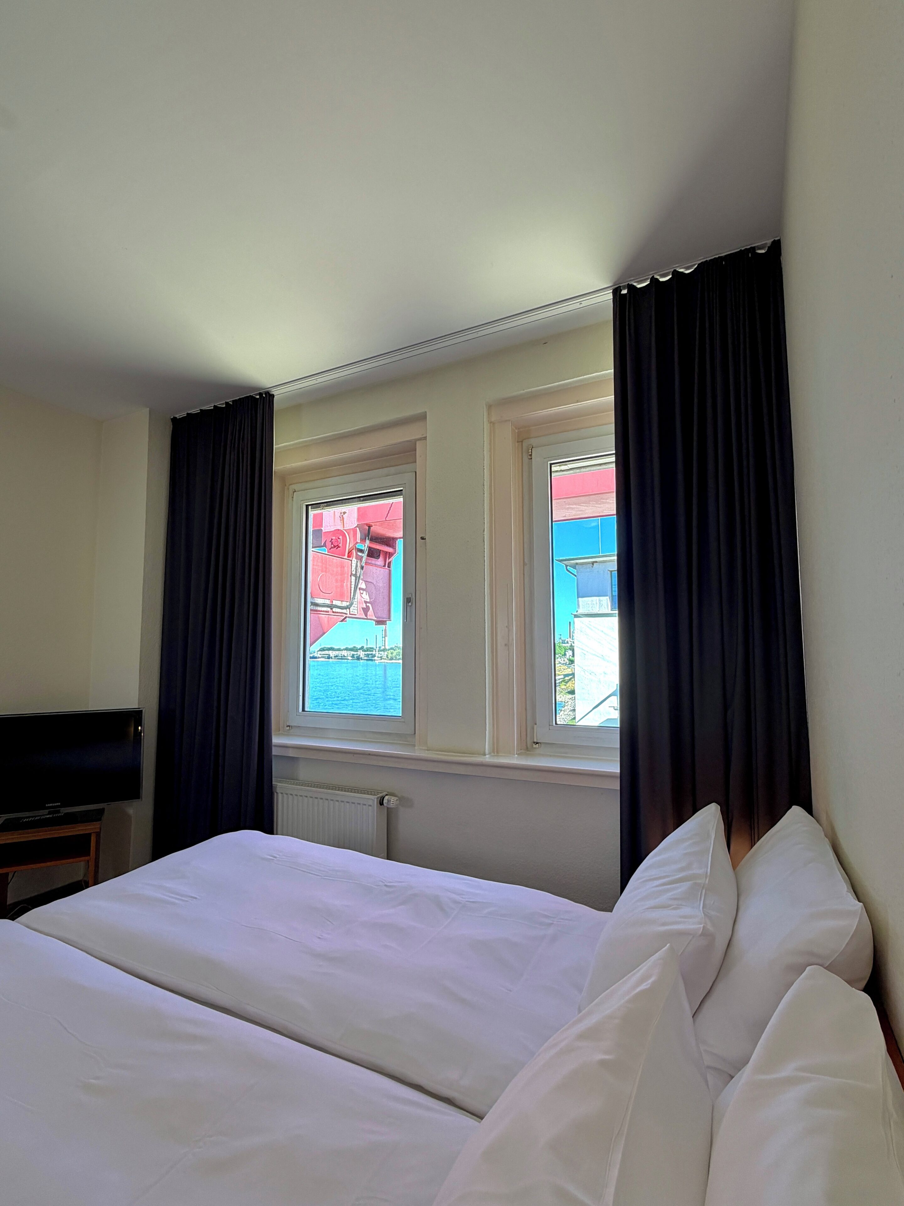 A hotel room with a bed, a television on a stand, and windows with curtains.