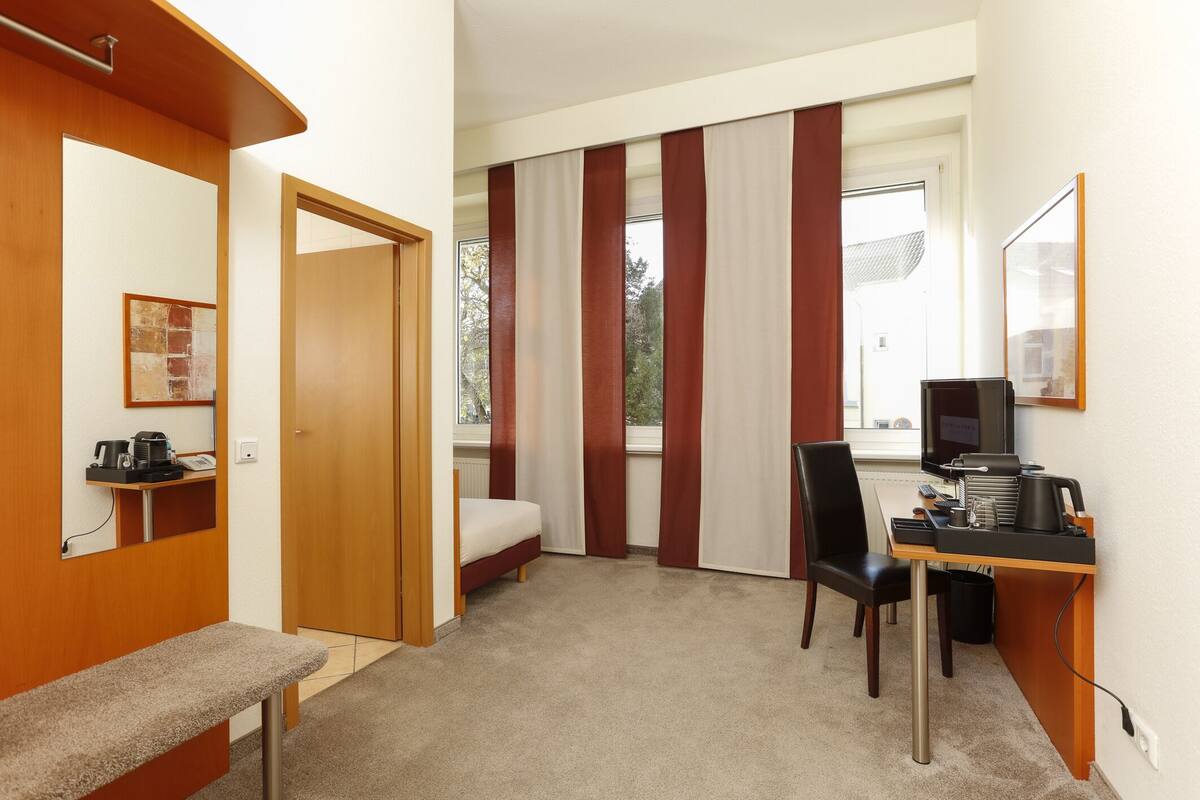 A hotel room with a bed, a desk with a computer, a chair, a bench, and a window with curtains.