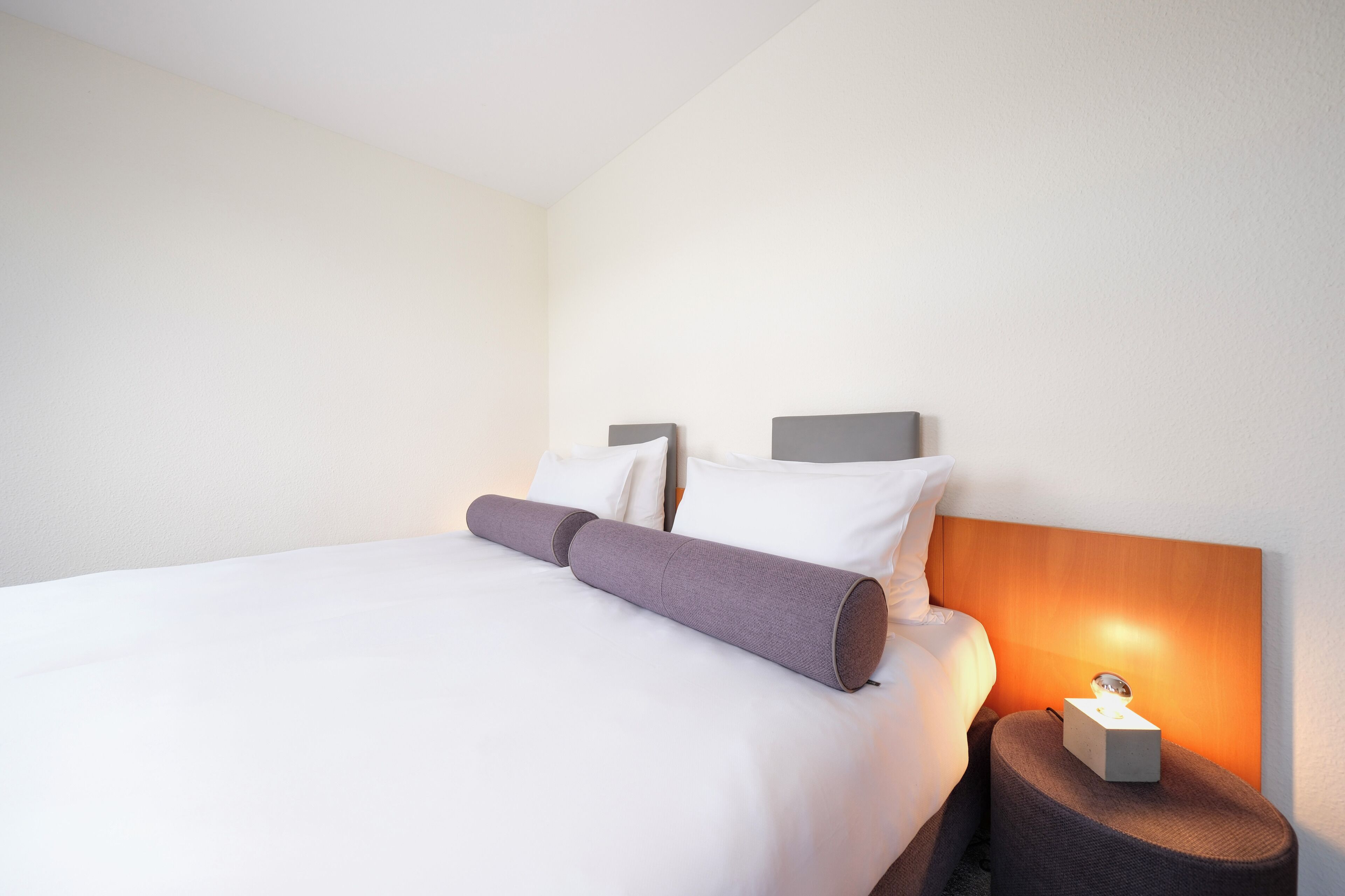 A neatly made bed with white sheets and a gray pillow, a bedside table with a lamp, and a wall-mounted headboard.