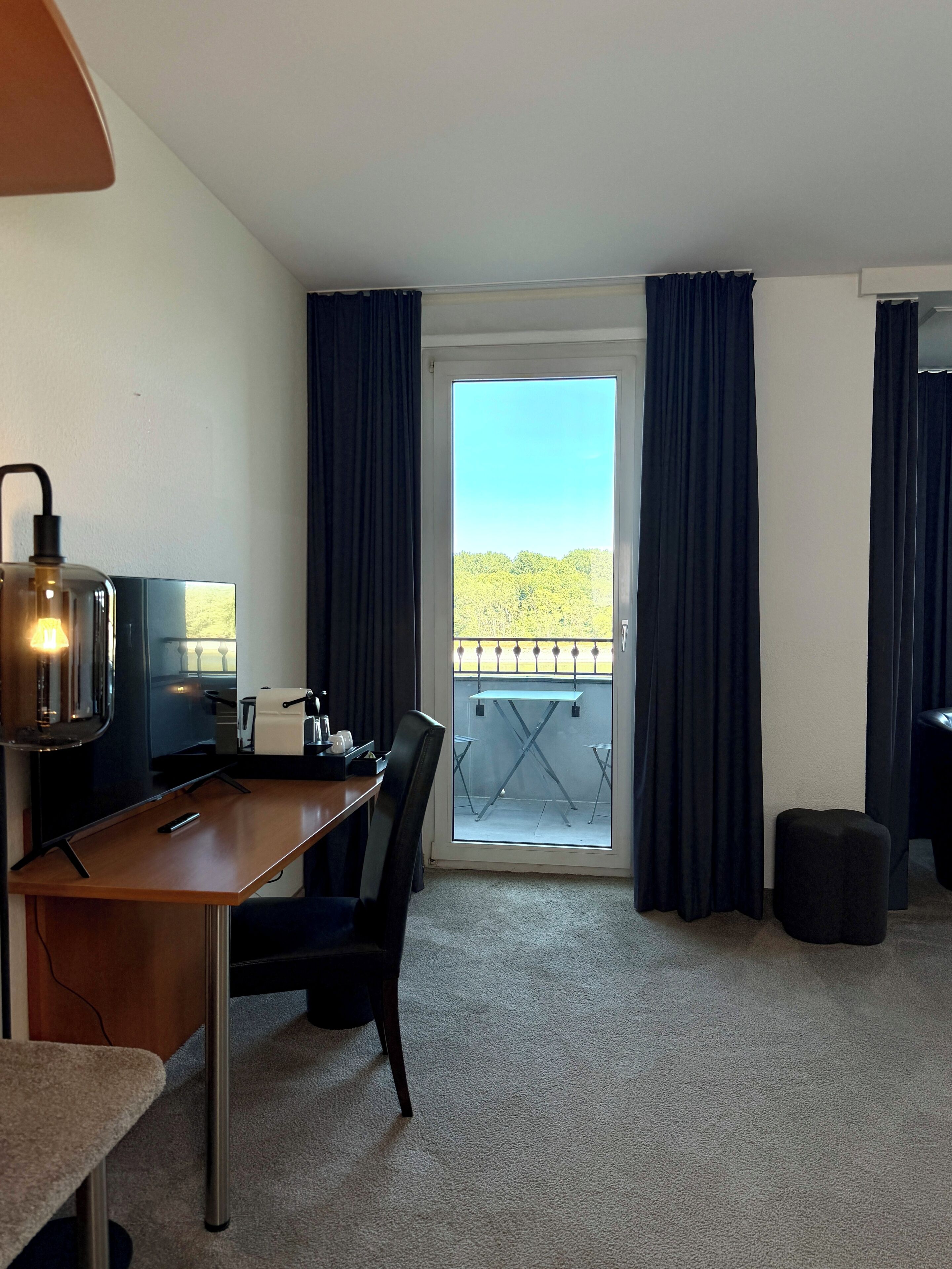 A hotel room with a desk, chair, and a view of a balcony with curtains.