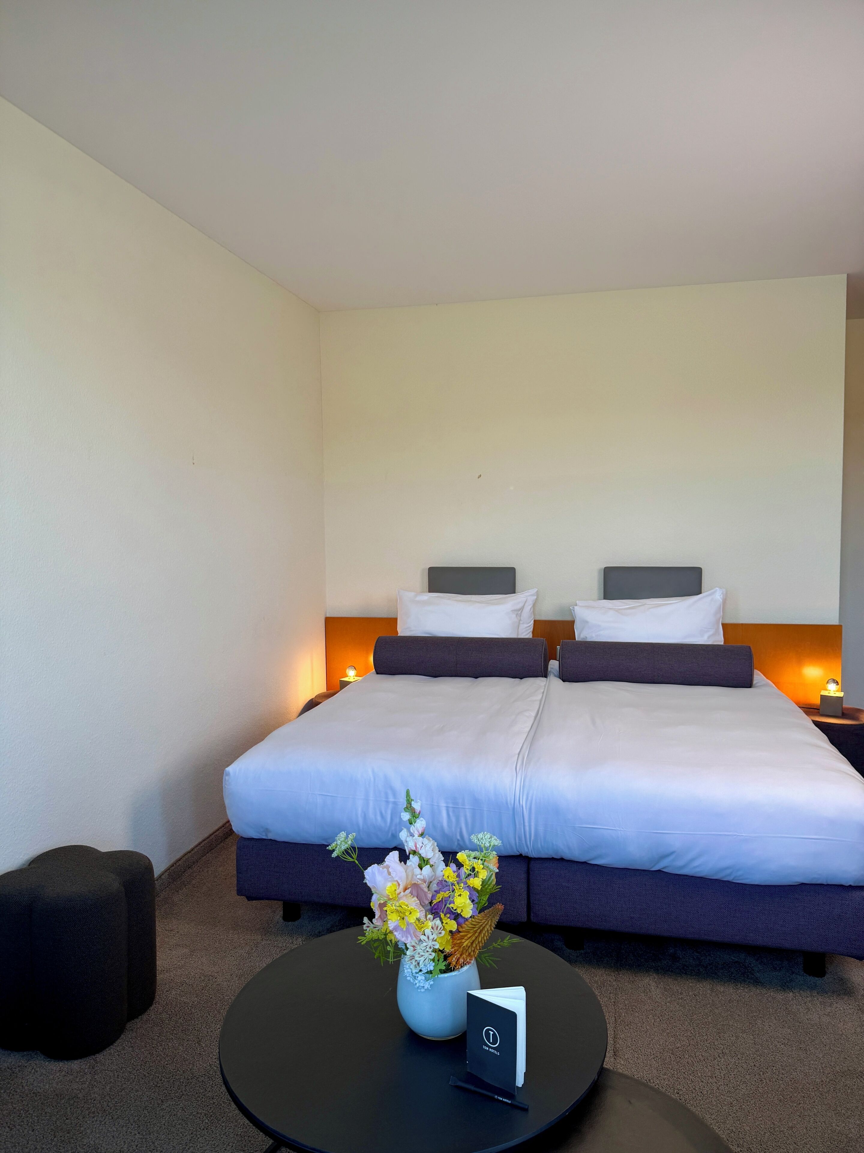 A hotel room with two beds, a small round table with a vase of flowers, and a nightstand with a lamp.