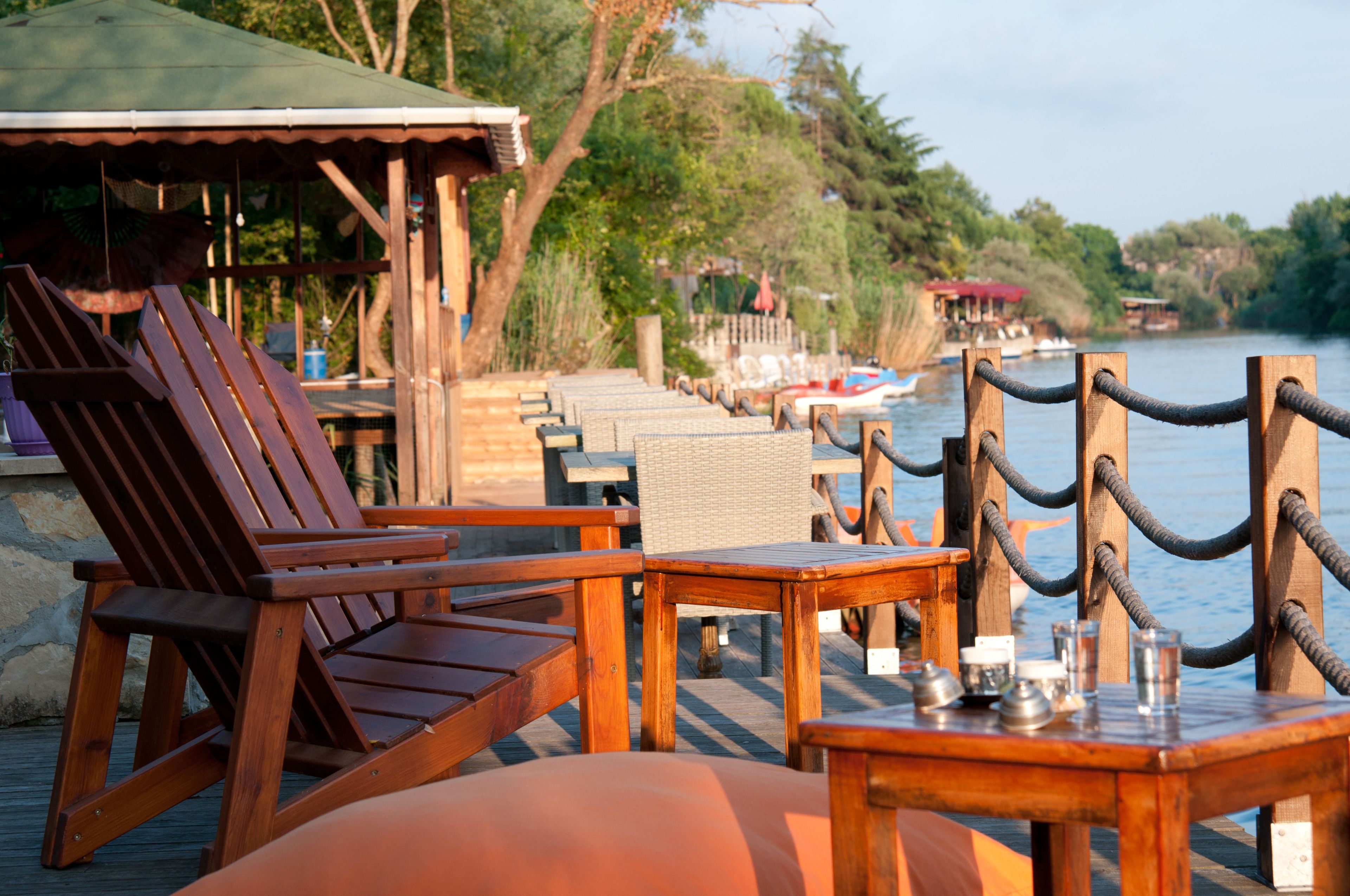A wooden deck with lounge chairs and tables by a lake, featuring a rope barrier and distant boats.