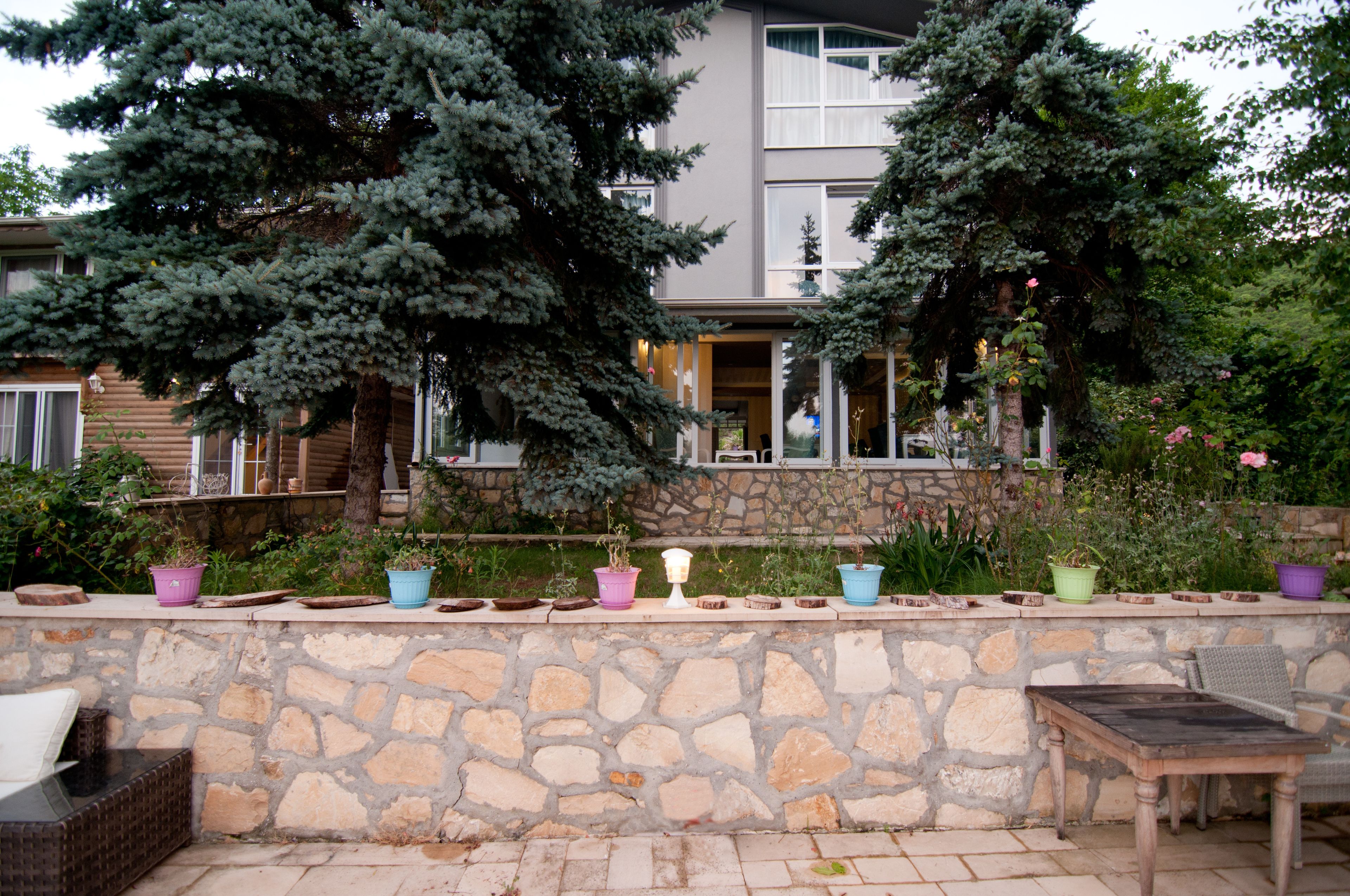 A stone wall with potted plants, a wicker chair, and a wooden table in a courtyard area.