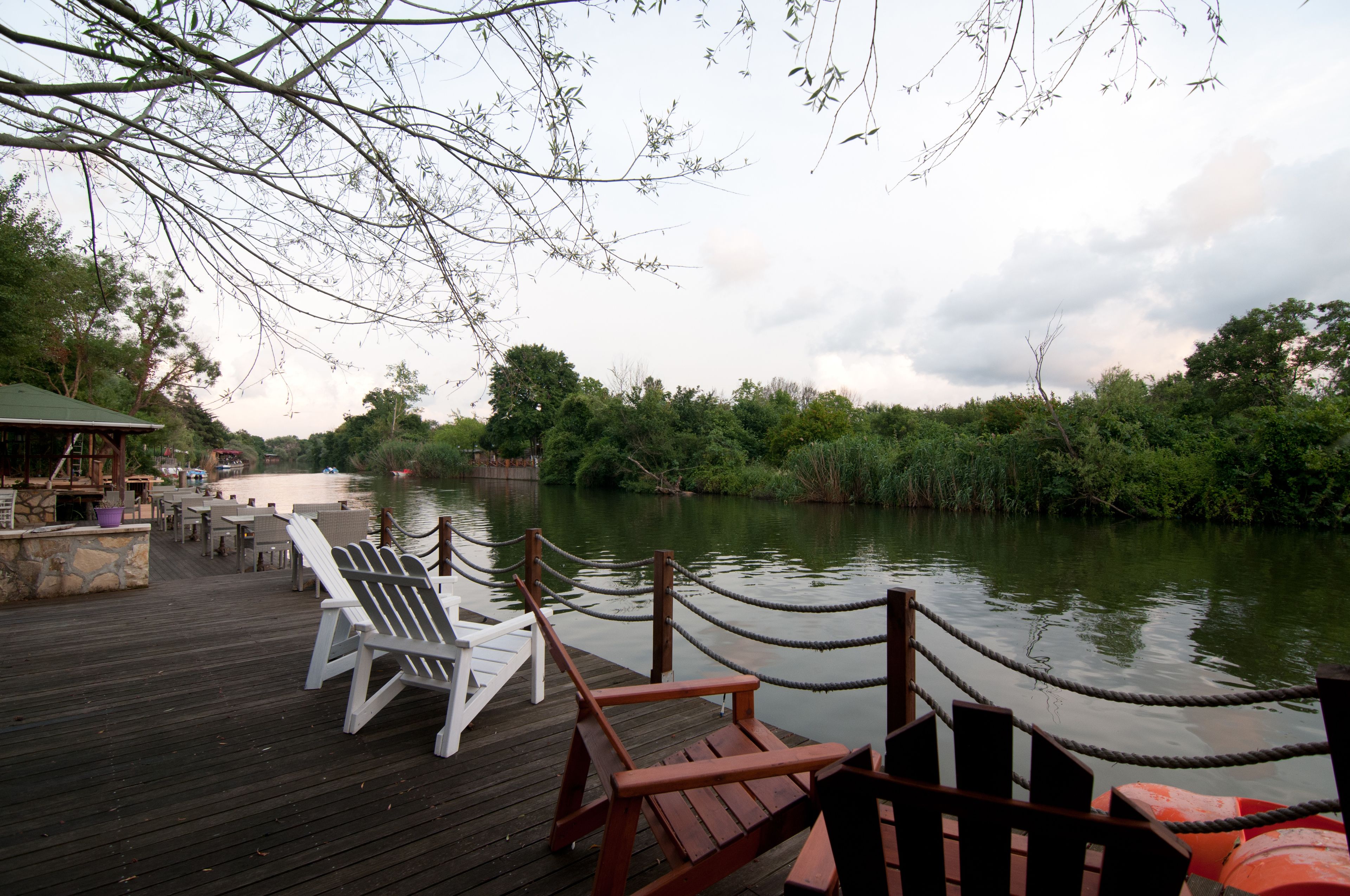 A wooden deck with chairs and tables overlooking a river, surrounded by lush greenery.