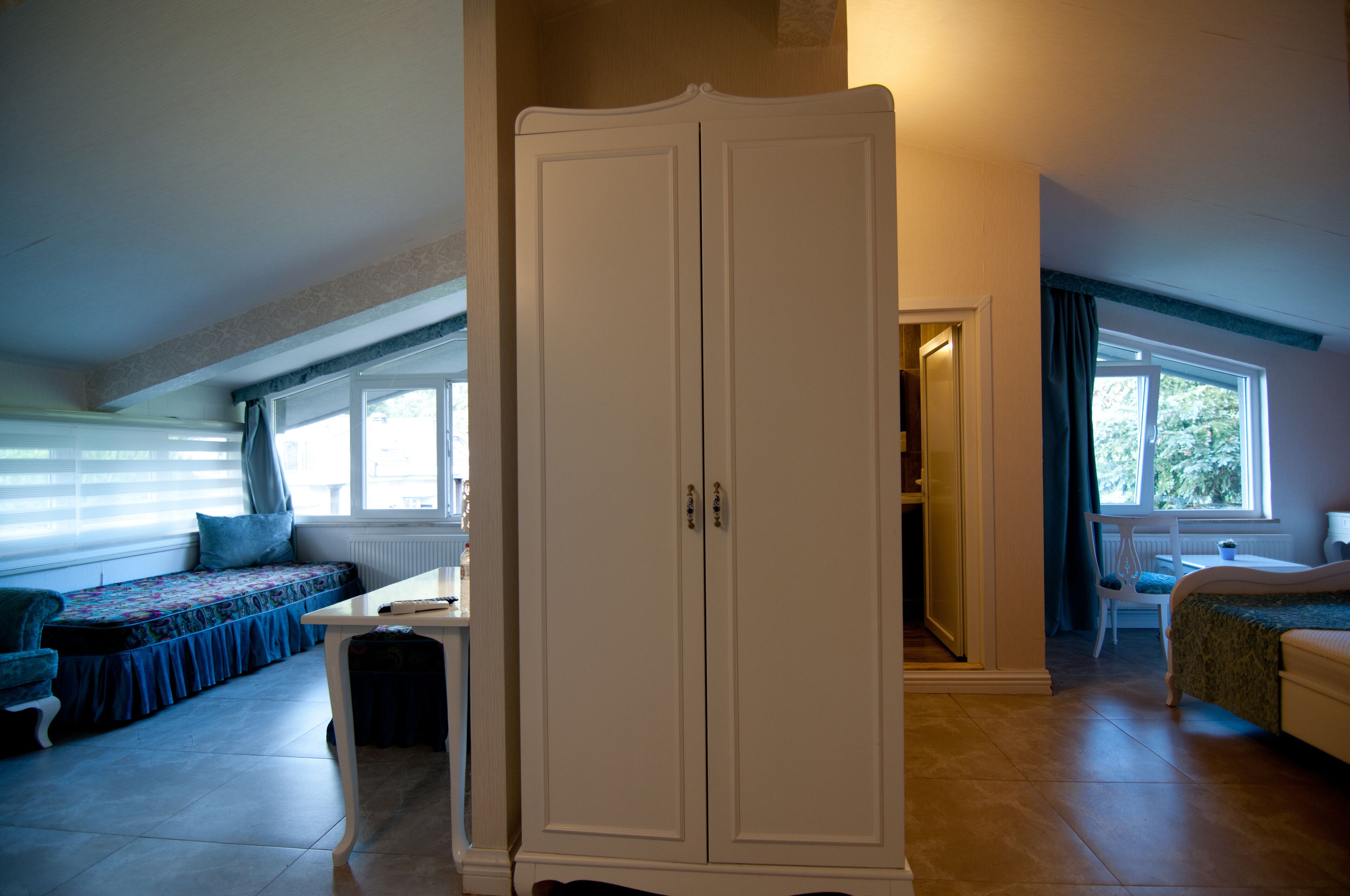 A room with a large white wardrobe, a small table, a bench with cushions, and a bed.