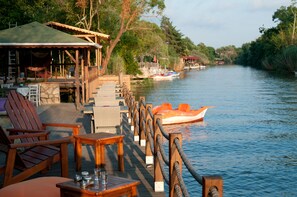 A wooden deck with chairs and tables, a boat on the water, and a building with a green roof in the background. - Ağva Nehir Evi Butik Hotel (Sile)