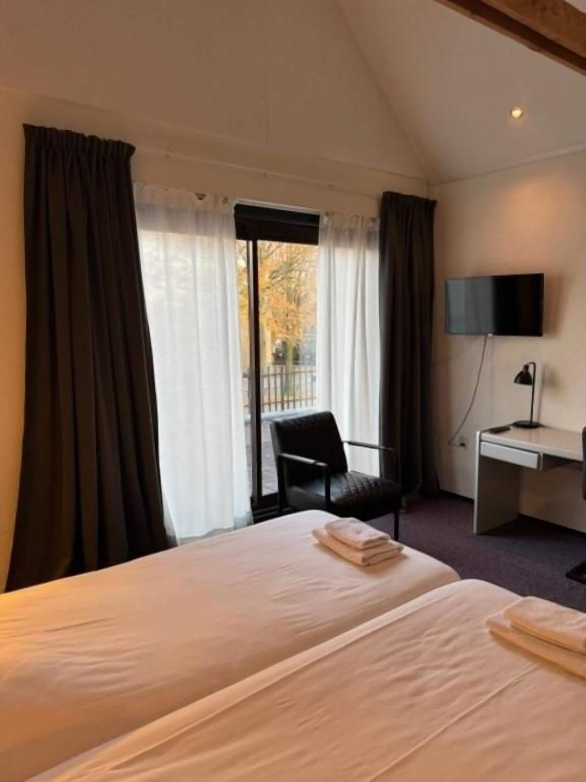Ett hotellrum med två sängar, ett skrivbord, en stol, en TV och ett fönster med gardiner.