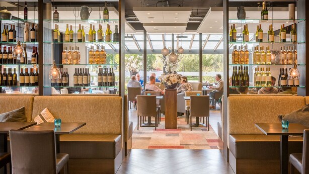 A modern bar with glass shelving displaying various bottles, a central seating area with tables and chairs, and a view through large windows. - Hotel Schimmel (Woudenberg)