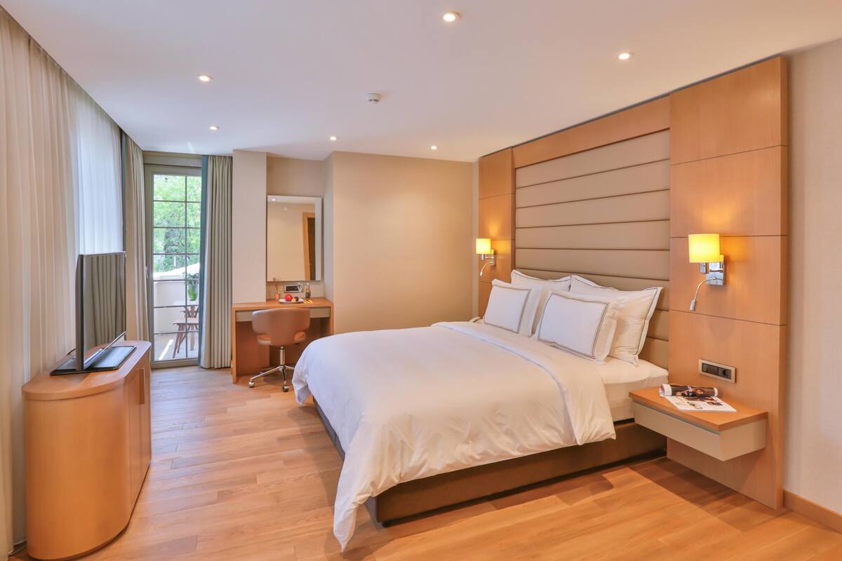 A modern hotel room with a large bed, bedside tables, a desk, and a window with curtains.