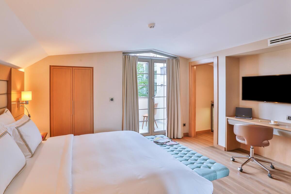 A hotel room with a large bed, a desk with a chair, a television, and a balcony with curtains.
