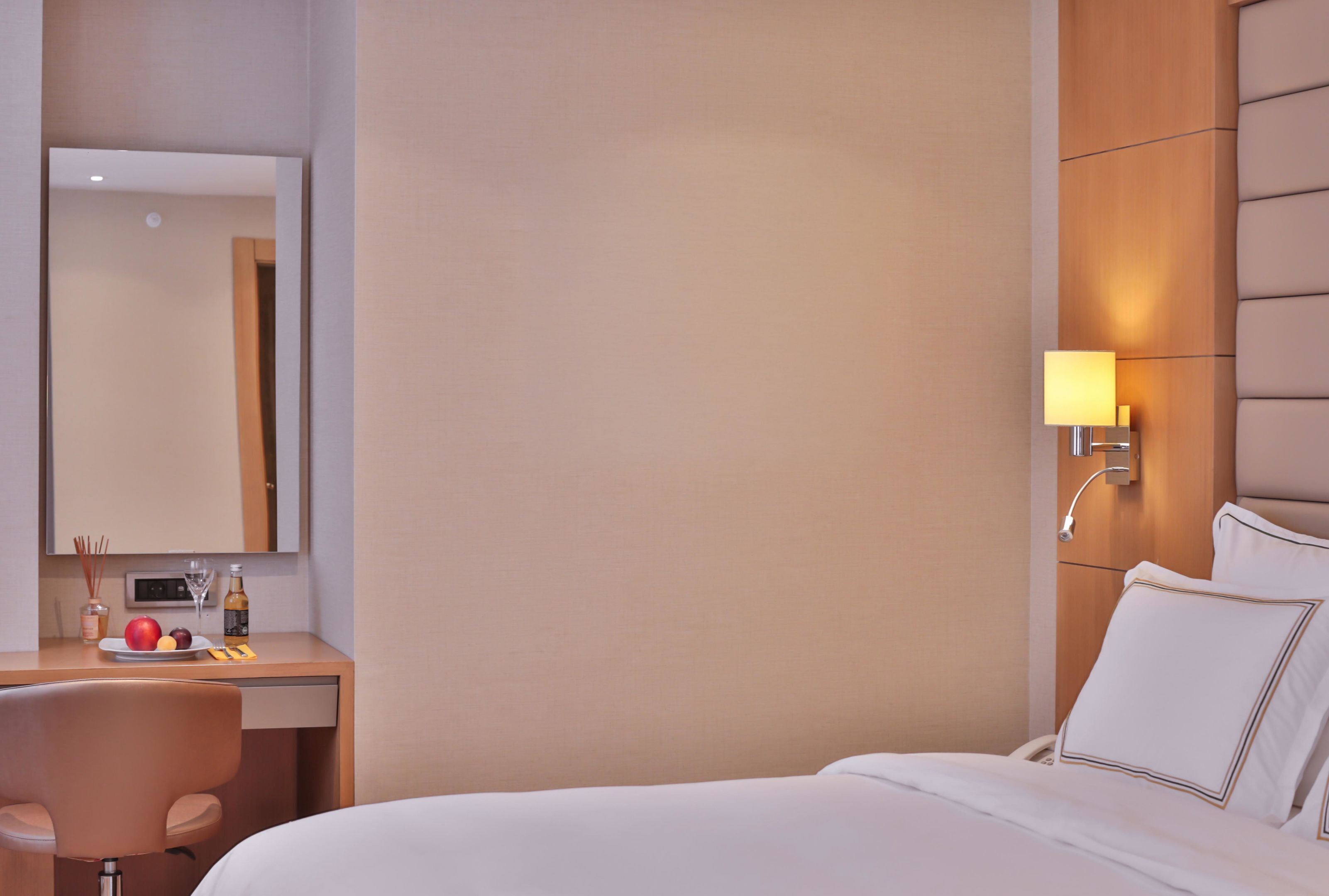 Ett hotellrum med en säng, ett skrivbord med en stol, en spegel, en vägglampa och ett litet bord med en flaska, ett glas och frukt.