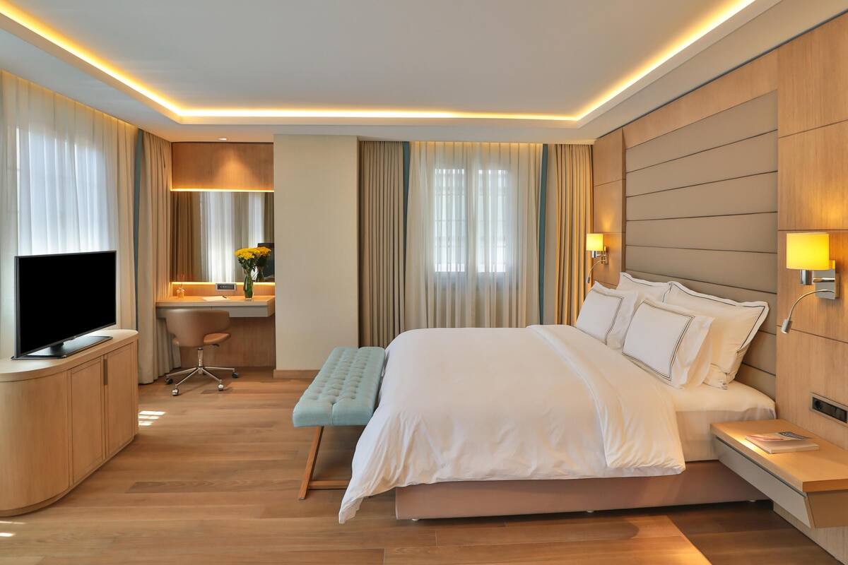 A hotel room with a bed, bedside table, TV, desk, chair, and a desk lamp.