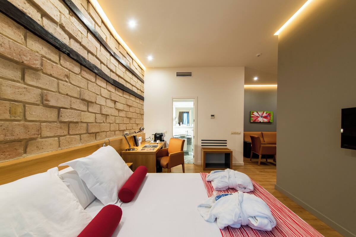 Hotelkamer met een bakstenen muur, een bed met witte lakens, een rood kussen, een rode deken en een rode badjas. Er is een bureau met een stoel, een kleine tafel en een aan de muur bevestigde televisie.