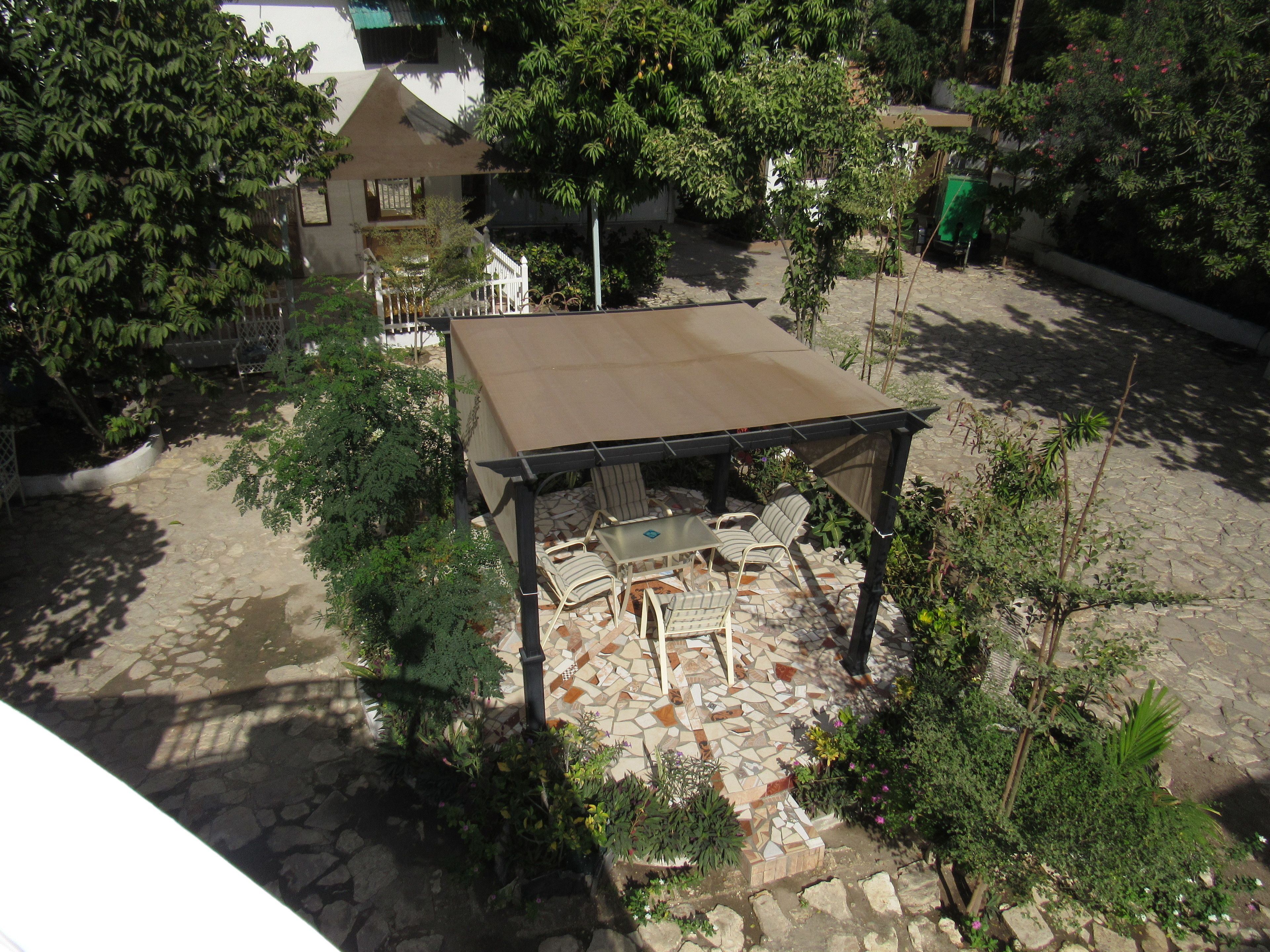 A covered outdoor seating area with chairs and a table, surrounded by greenery and a stone-tiled patio.