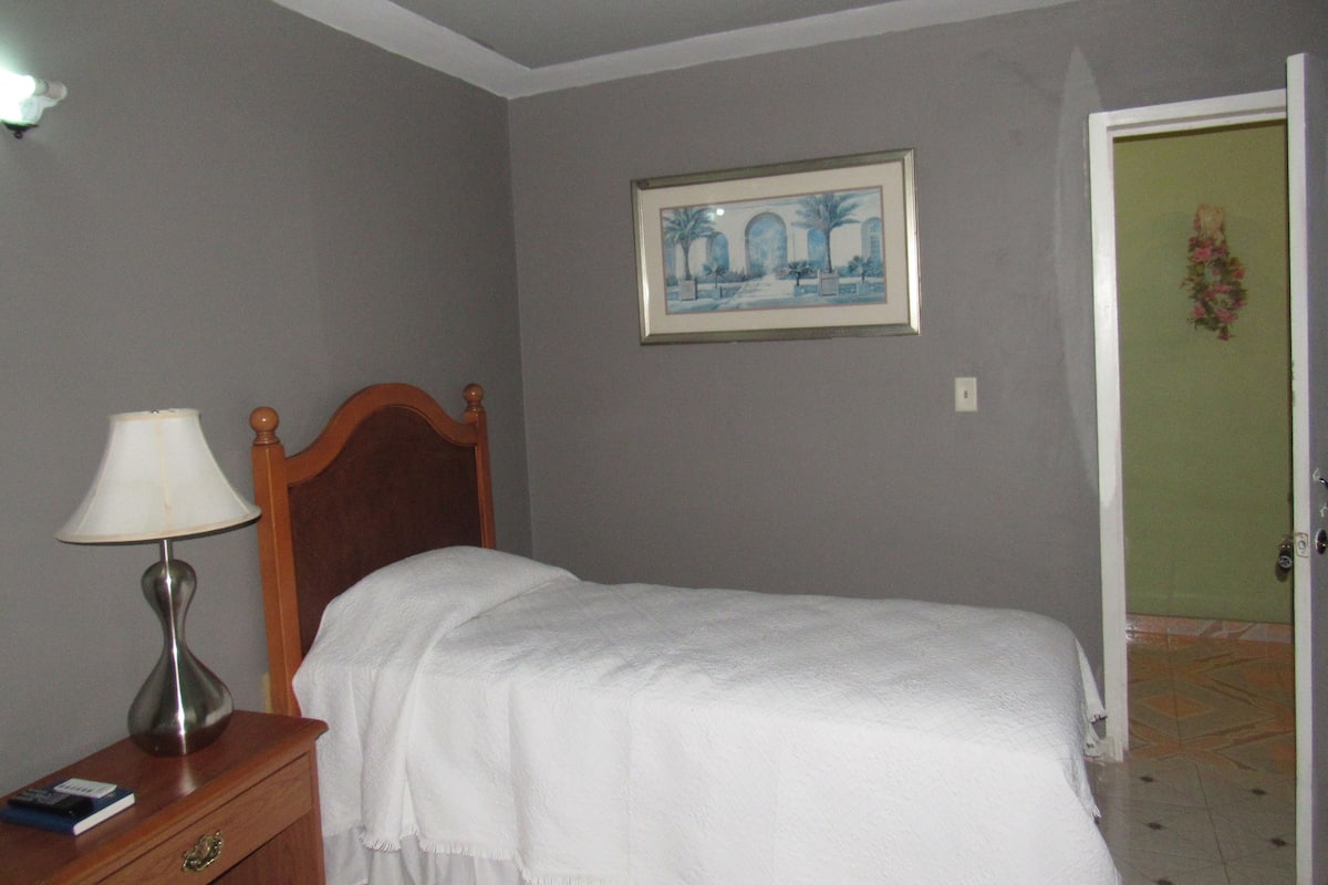 A bedroom with a bed, a nightstand with a lamp, a framed picture on the wall, and a door to the right.