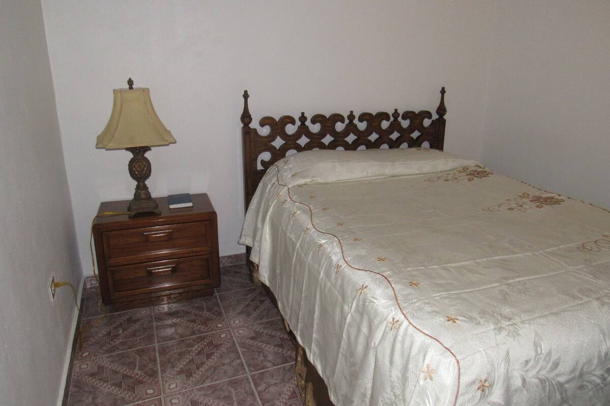 A bedroom with a wooden bed frame, a bedside table with a lamp, and patterned floor tiles.