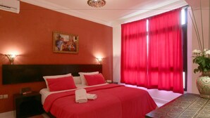A bedroom with a large bed, red curtains, a painting on the wall, and a vase with flowers on a table. - Hotel Tafoukt (Marrakech)