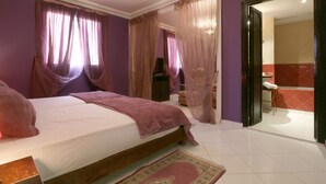 A bedroom with a bed, curtains, a television, and a bathroom visible through an open door. - Hotel Tafoukt (Marrakech)
