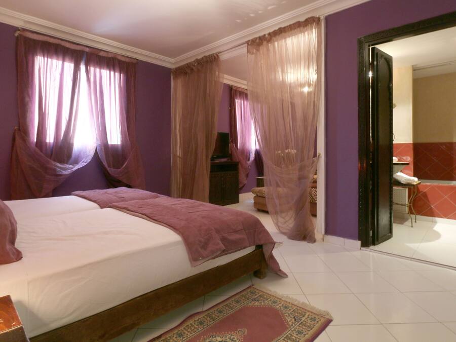 Une chambre à coucher avec un lit, des rideaux, un téléviseur et une salle de bain visible à travers une porte ouverte.