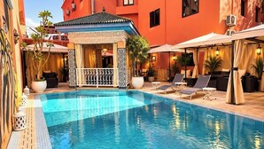 A swimming pool with lounge chairs and umbrellas, surrounded by a building with a tiled floor and a covered seating area. - Hotel Tafoukt (Marrakech)
