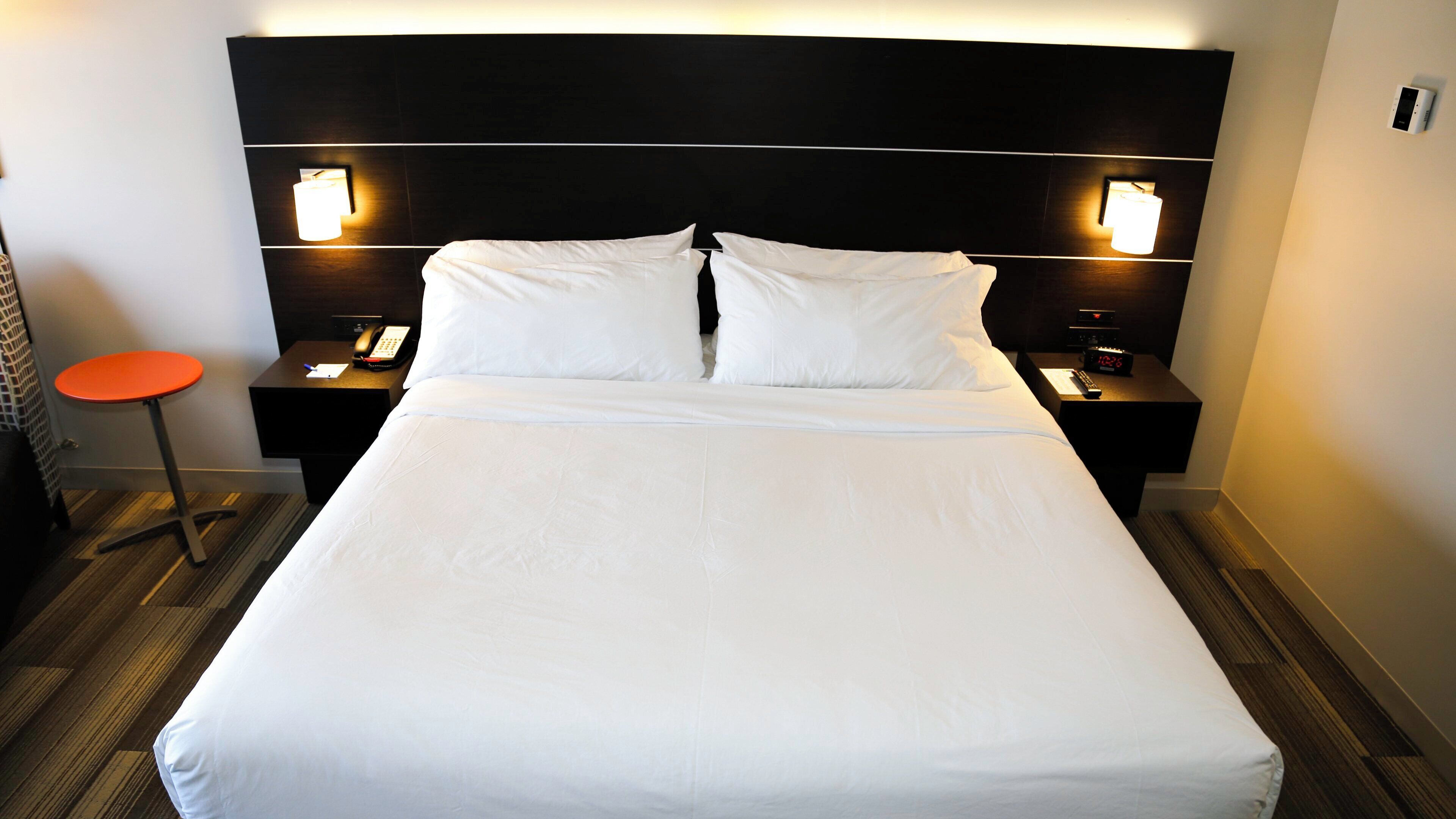 Ein Hotelzimmer mit einem großen Bett, zwei Nachttischen, einer Ablage, einem roten Hocker und Wandleuchten.