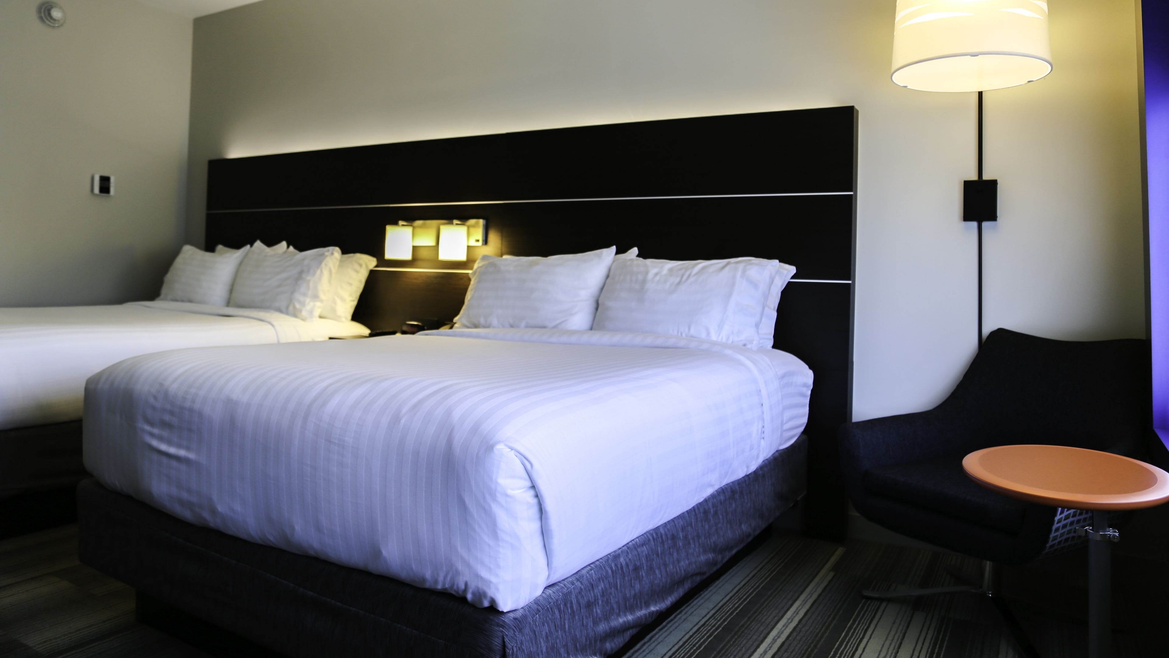 A hotel room with a large bed, a smaller bed, a nightstand with lamps, a chair, and a table.