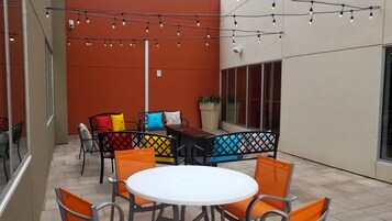 Un patio exterior con una mesa redonda y sillas naranjas, una cerca de metal negro y luces colgantes.