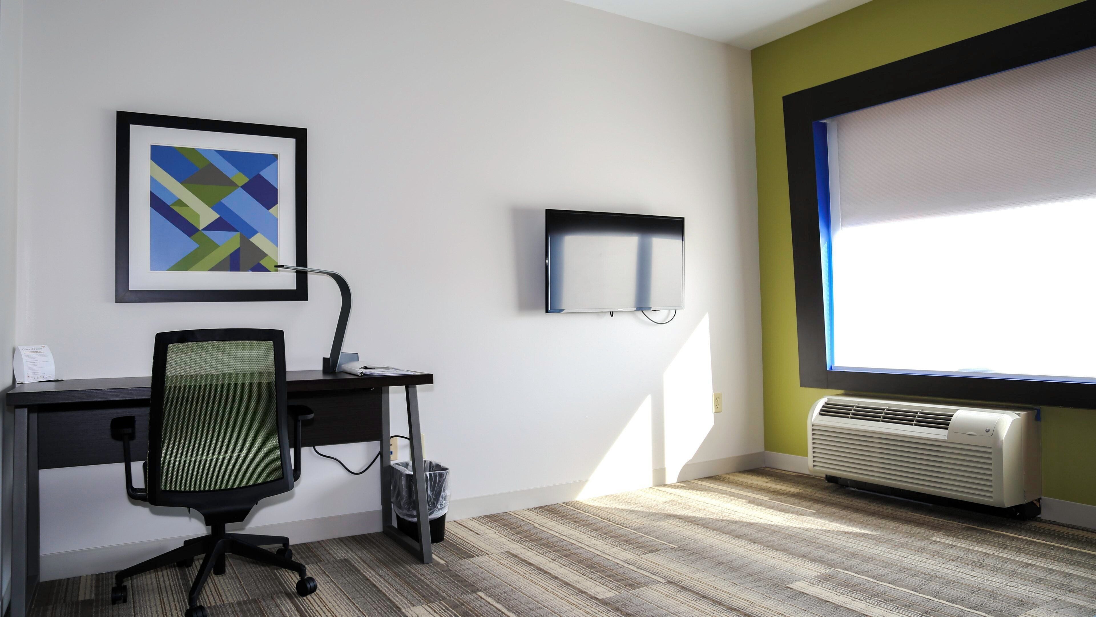 A modern office space with a desk, chair, framed artwork, a flat-screen monitor, and a window with blinds.