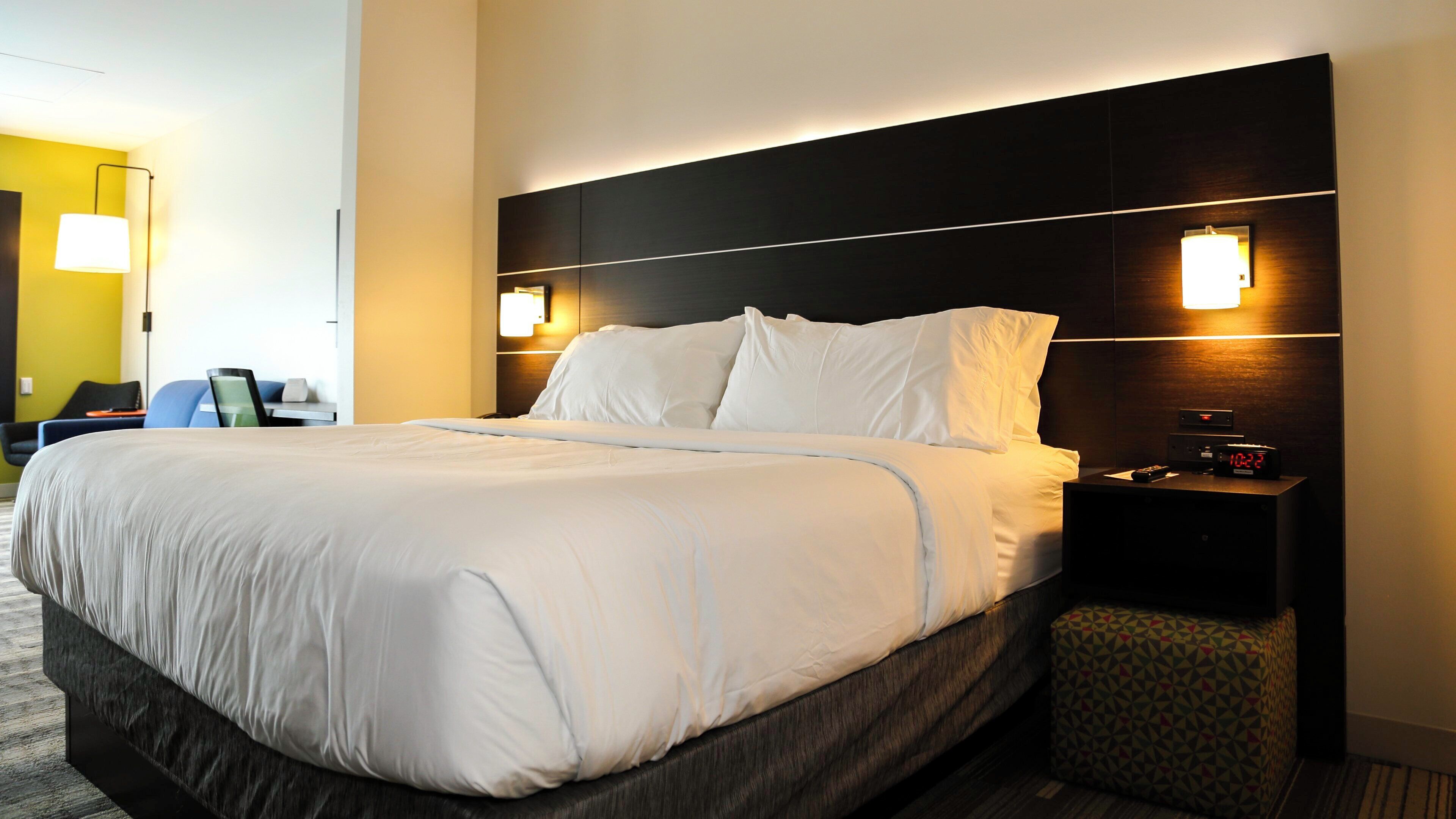 A hotel room with a large bed, a bedside table with a clock, and a wall-mounted lamp.