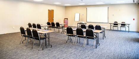 Un salón de conferencias con una pizarra, proyector y varias mesas con sillas.