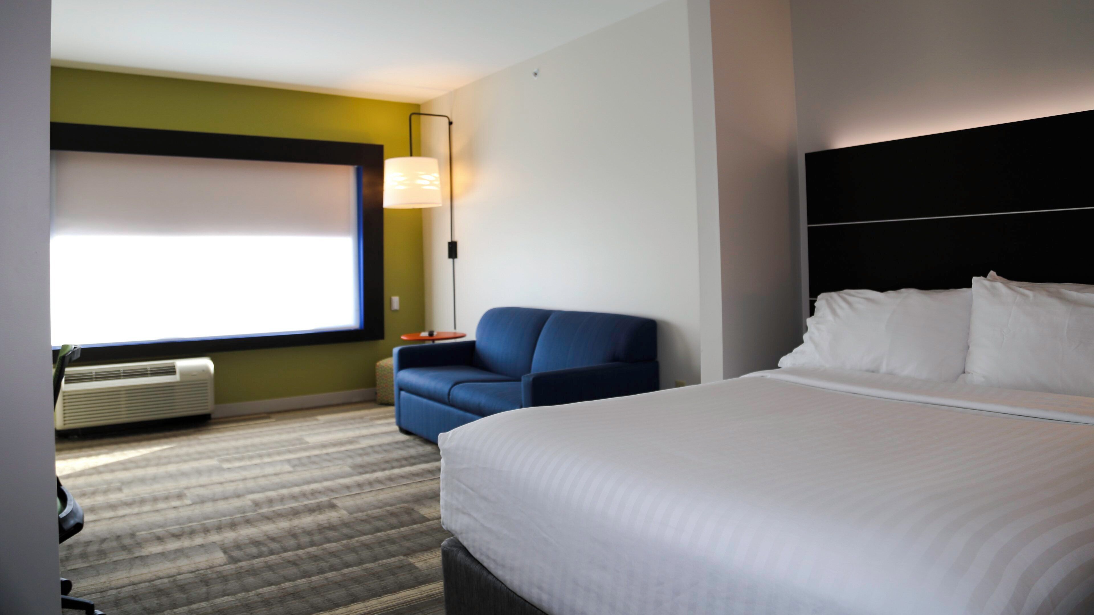 A hotel room with a bed, a blue armchair, a desk, a lamp, and a window.