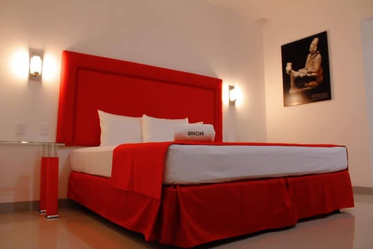 Habitación de hotel con cabecera roja, ropa de cama blanca y lámpara de pared.