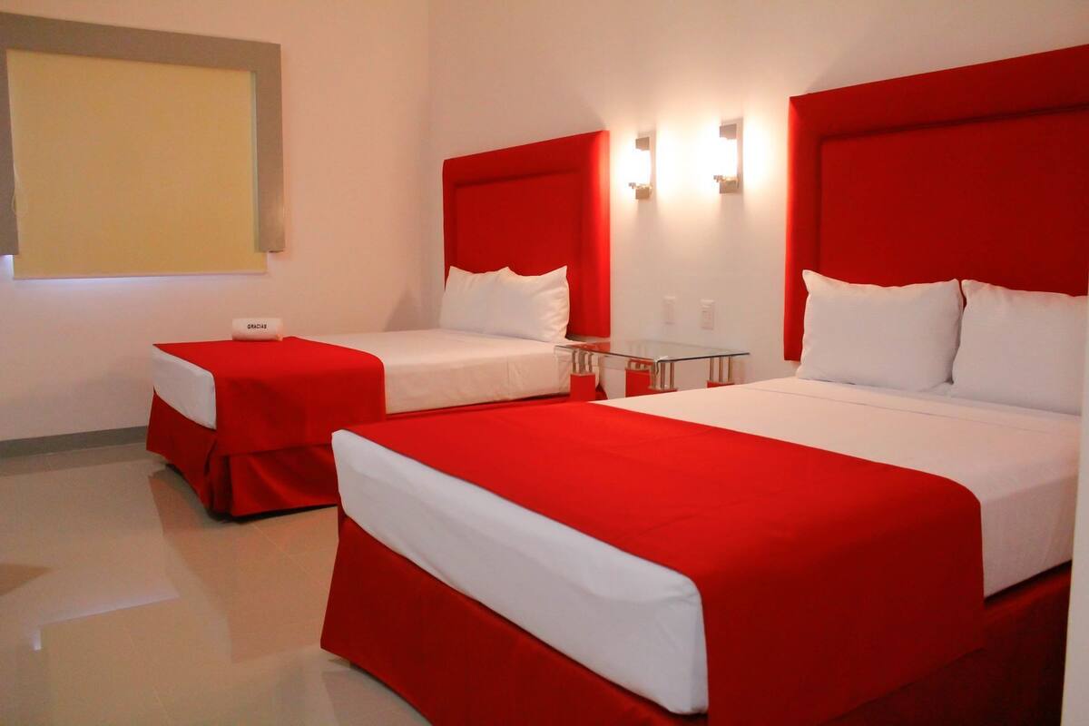 Habitación de hotel con dos camas, cada una con ropa de cama roja y blanca, una mesita pequeña y una lámpara fijada a la pared.