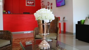 A reception area with a red wall, a 'Zar Hotel' sign, a glass table with a silver vase of white flowers, wicker chairs, and a television on the wall.