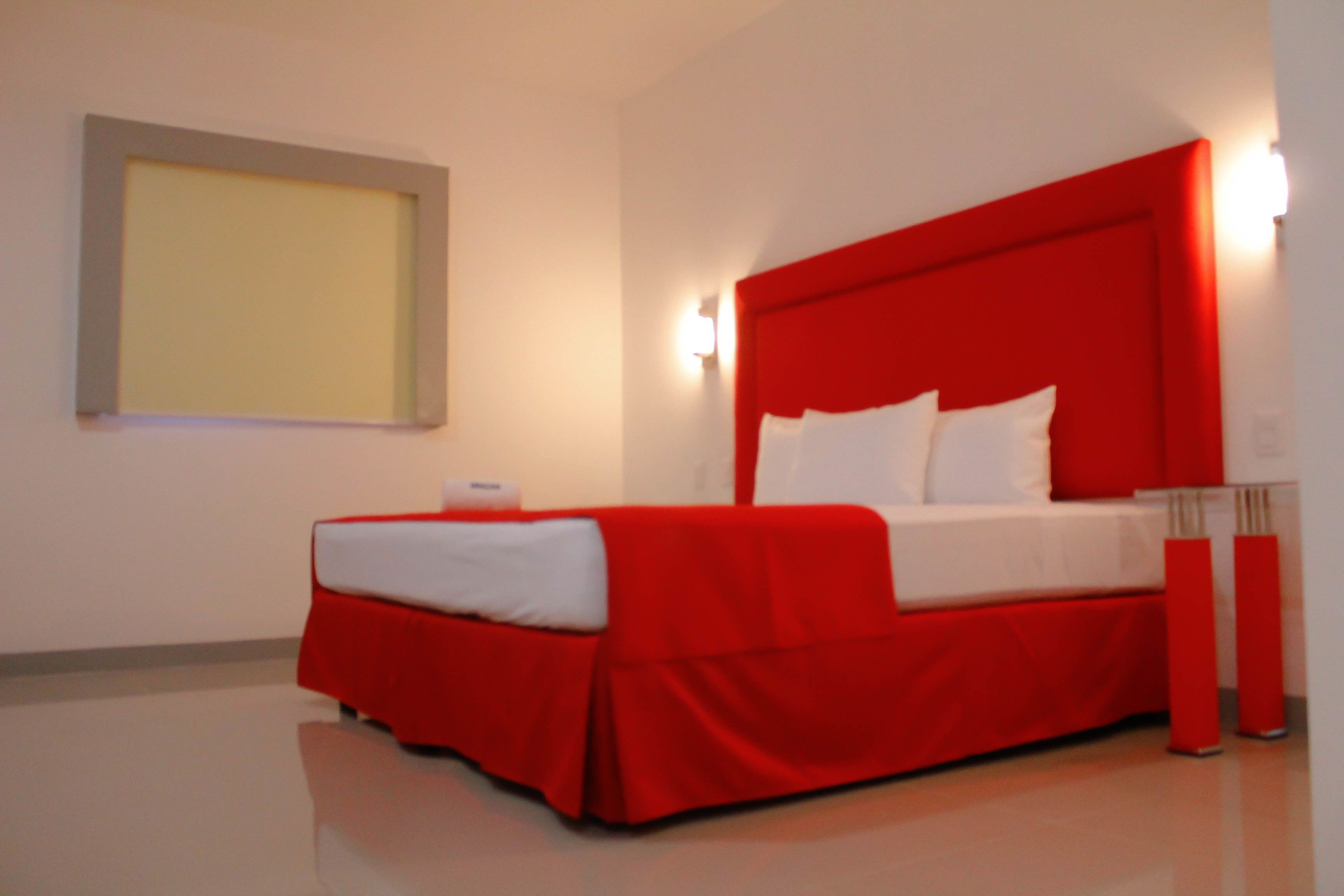 A red bed with white bedding in a room with a plain wall and a light switch.