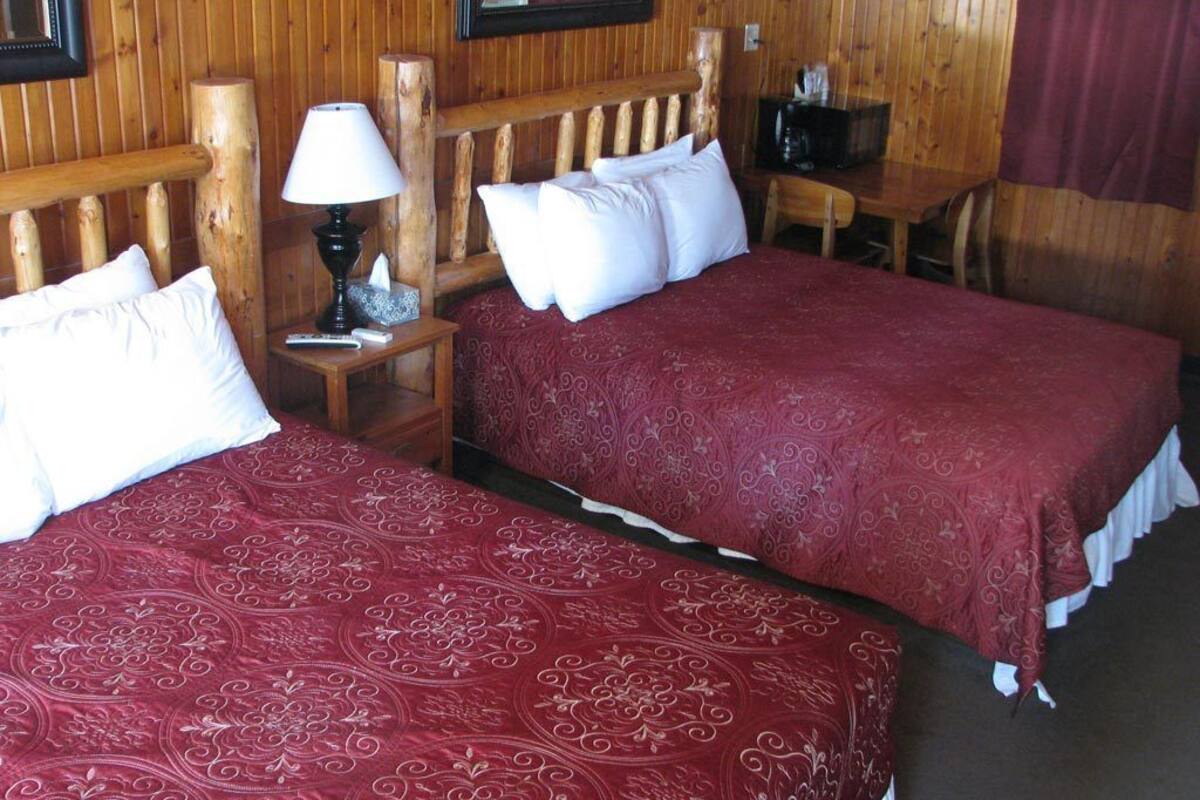 A room with two beds, wooden headboards, a nightstand with a lamp, a small table with a chair, and a television.