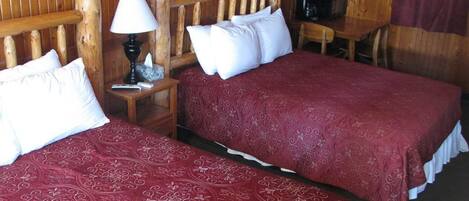 A room with two beds, wooden headboards, a nightstand with a lamp, a small table with a chair, and a television.