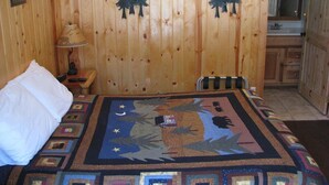 A wooden cabin room with a bed, a nightstand, a lamp, and a bathroom visible through an open door. - Evergreen Motel (Coram)