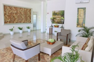 Un vestíbulo amplio con un cuadro abstracto de gran tamaño, varias plantas en macetas, una zona de estar con sofá y sillones, y un estante decorativo con jarrones y una cesta.