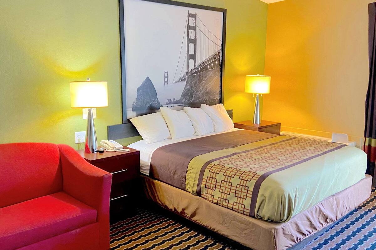 A hotel room with a bed, two bedside lamps, a red armchair, a framed picture of a bridge, and a patterned rug.
