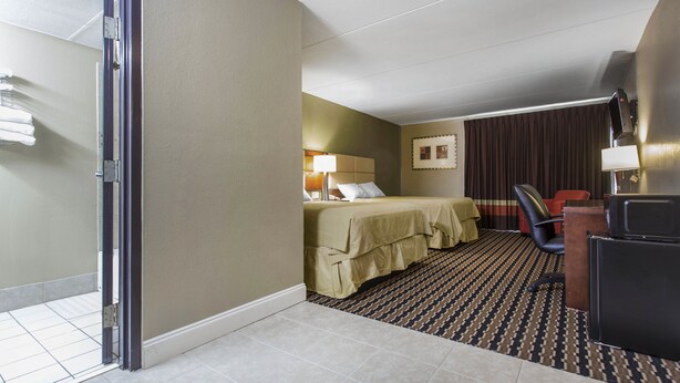 Quarto de hotel com cama, escrivaninha, cadeira, luminária e janela com cortinas.