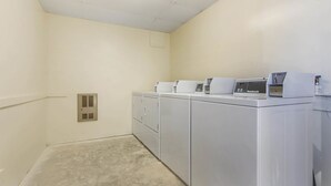 Uma lavanderia com uma fileira de eletrodomésticos brancos, incluindo máquina de lavar e secadora frontais, e um painel de controle no topo.
