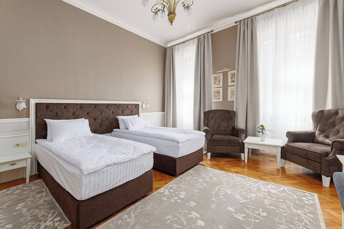 A hotel room with two beds, a sitting area with a chair, a small table, and a chandelier.