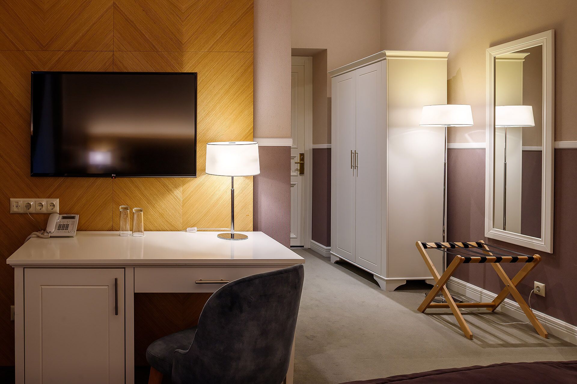 A hotel room with a flat-screen TV, a desk with a telephone, a chair, a mirror, and a wardrobe.