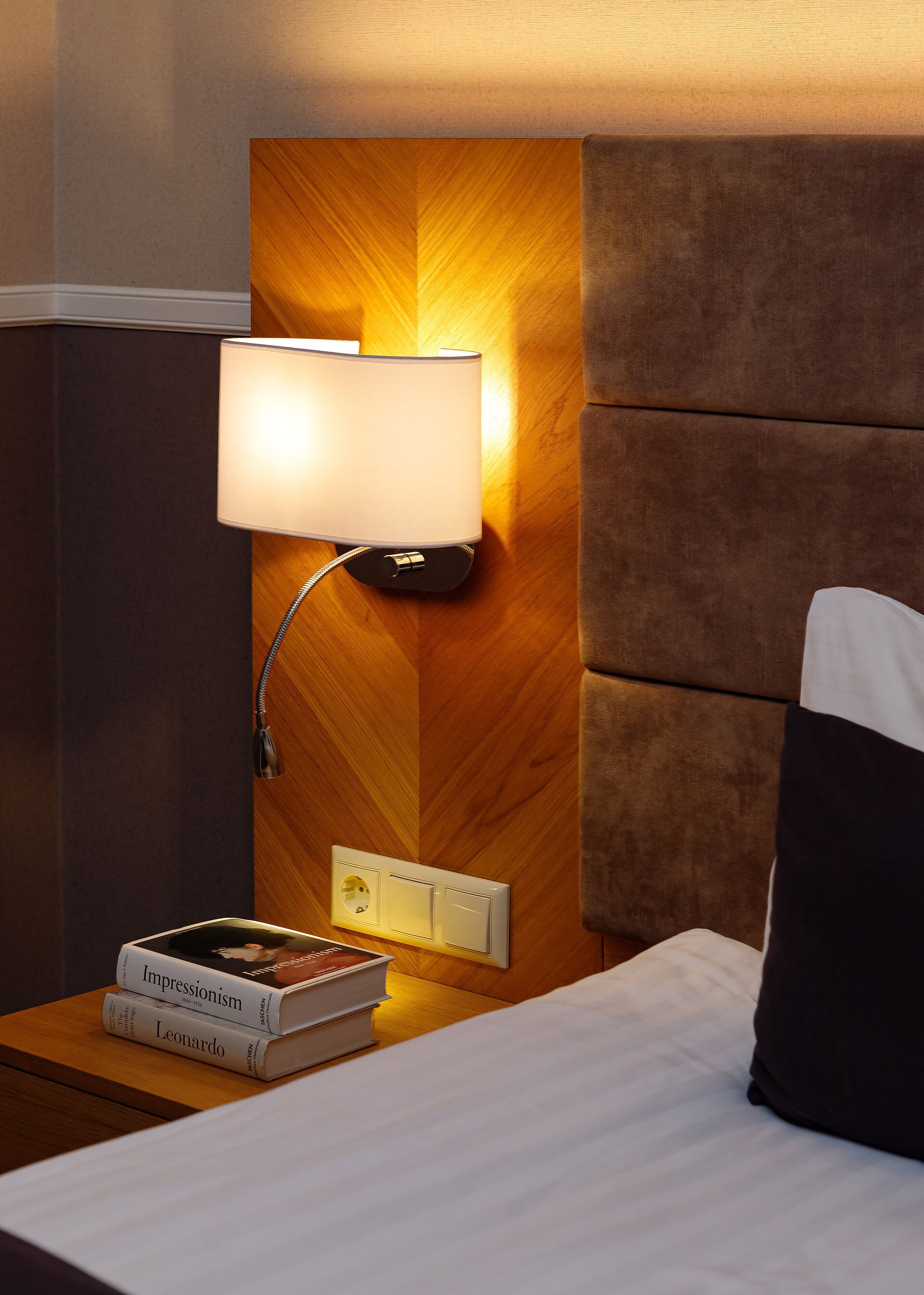 A neatly made bed with a pillow, a bedside lamp, and a stack of books on a wooden nightstand.